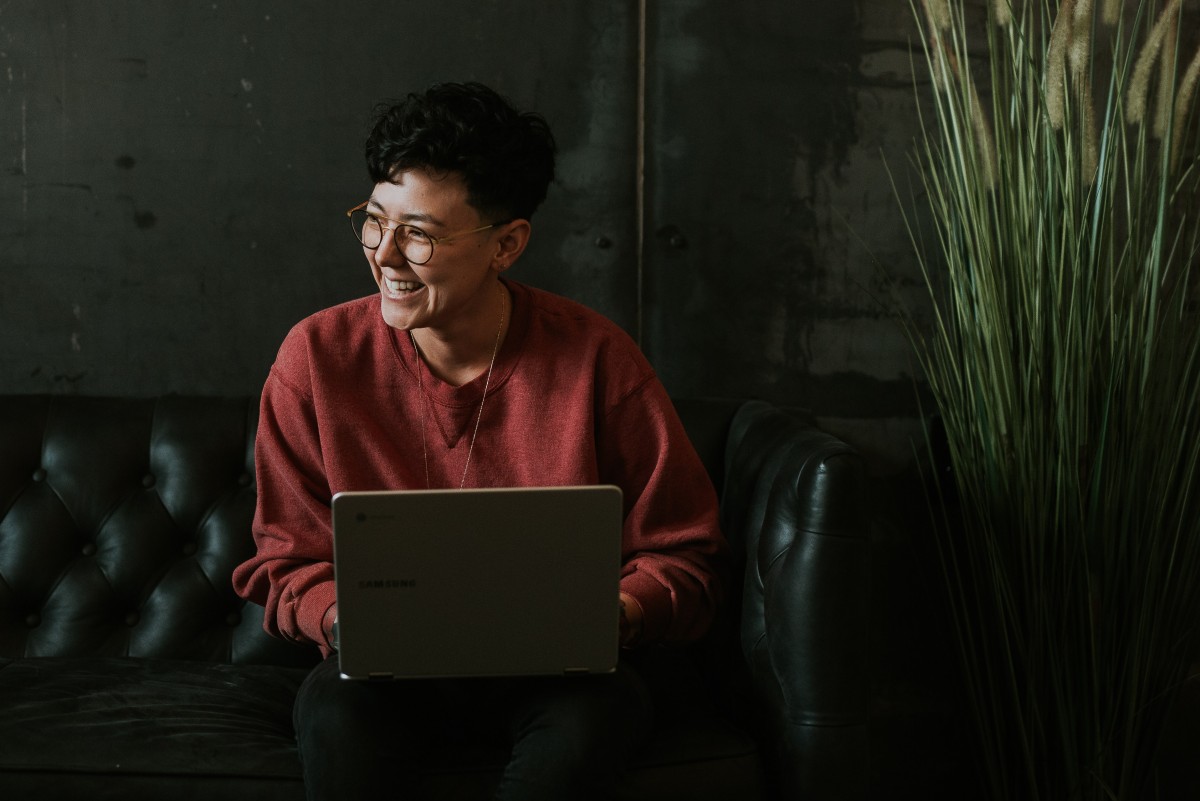 A person sitting on a black couch with a laptop is looking left and smiling.